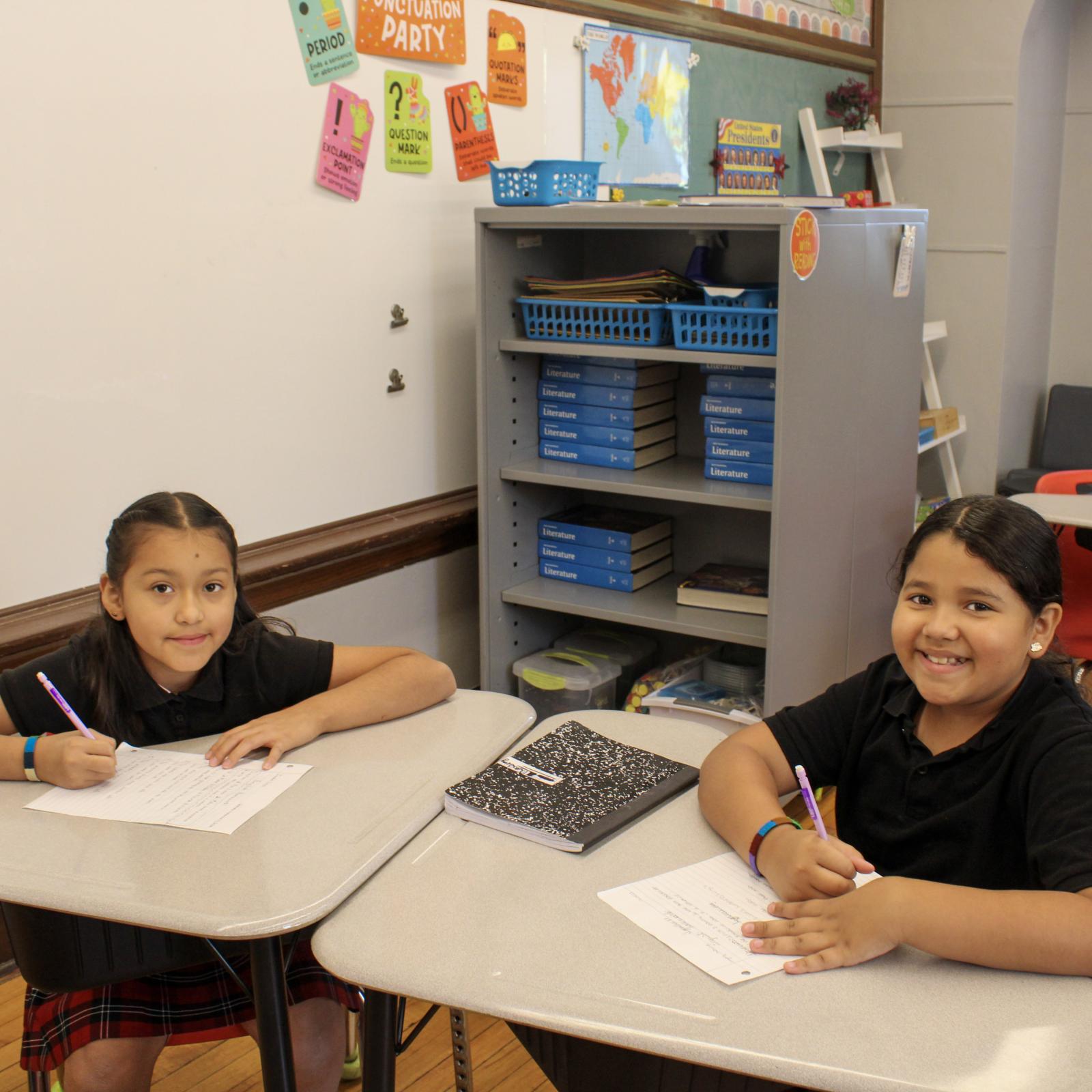 Two students at desks smiling.